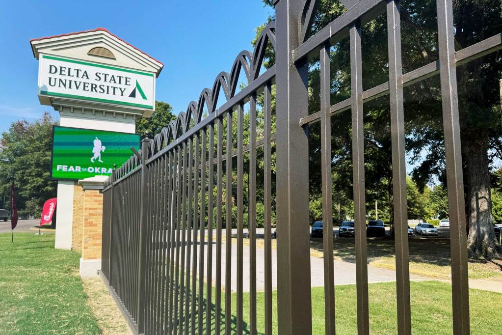 A iron fence leads to a sign in the distance that reads Delta State University