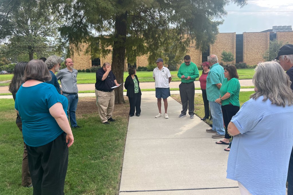 People stand outside of a brick building in the grass in a loose circle