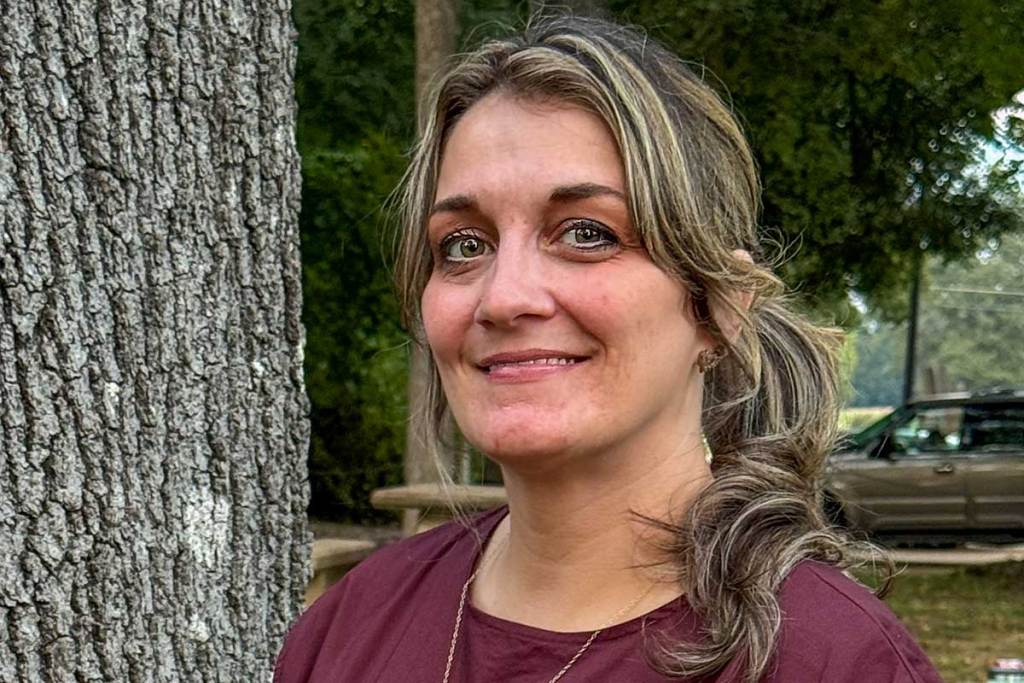 A woman in a burgundy blouse stands near a tree, smiling slightly.
