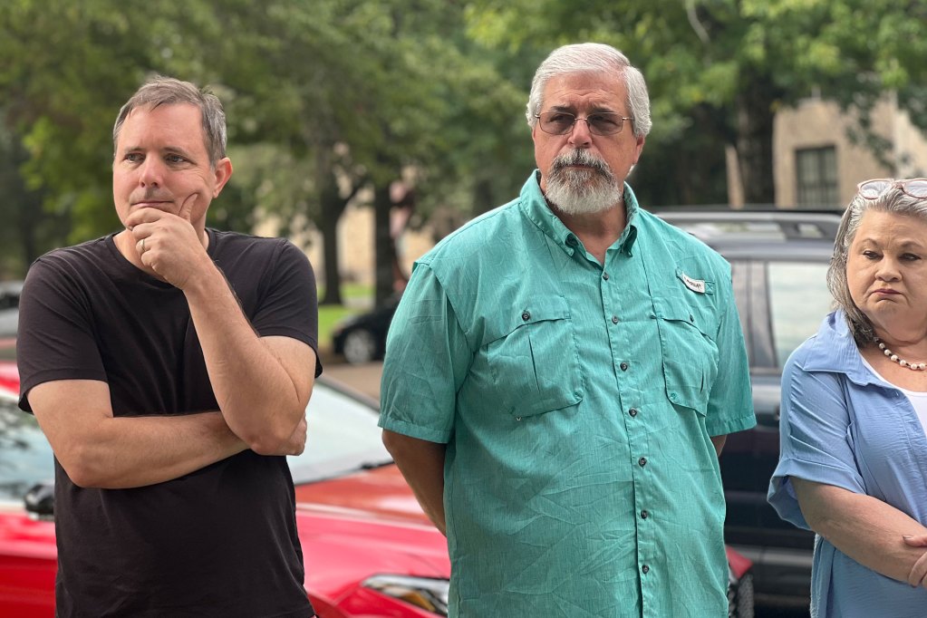 Three people stand outside in front of parked cars
