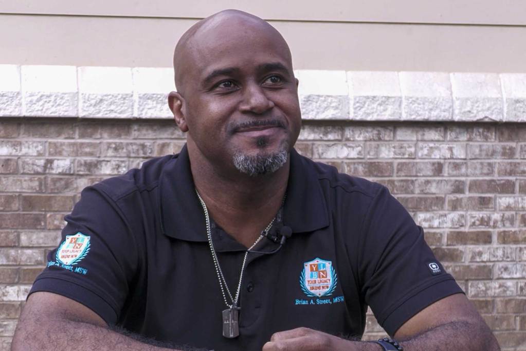 A man wearing a black polo sits at a picnic table in front of a brick wall.