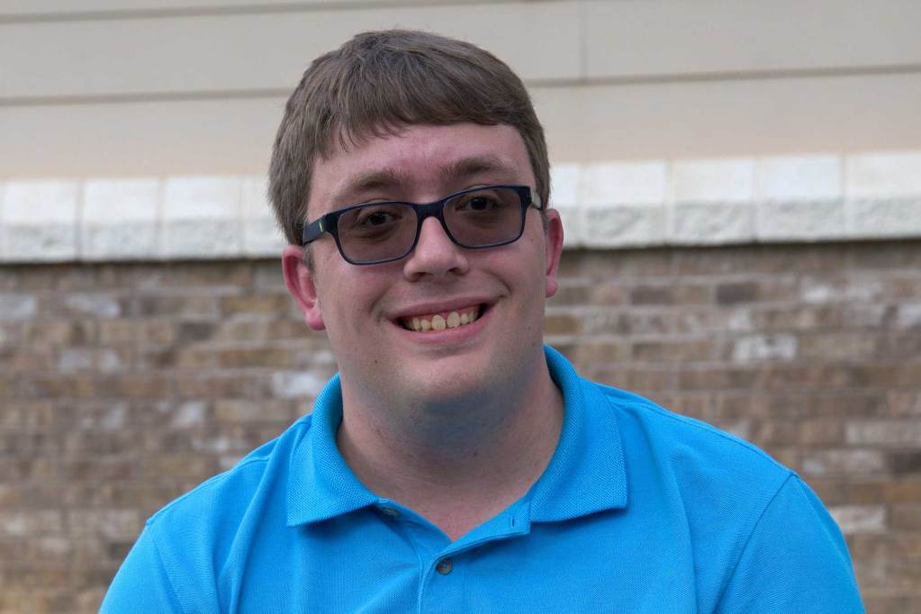 A man wearing a blue shirt smiles while sitting at a table outdoors.