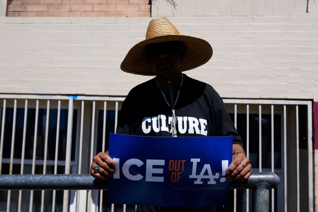 A person with their face shaded by a large brim had holds a sign that reads 'ICE OUT OF LA'