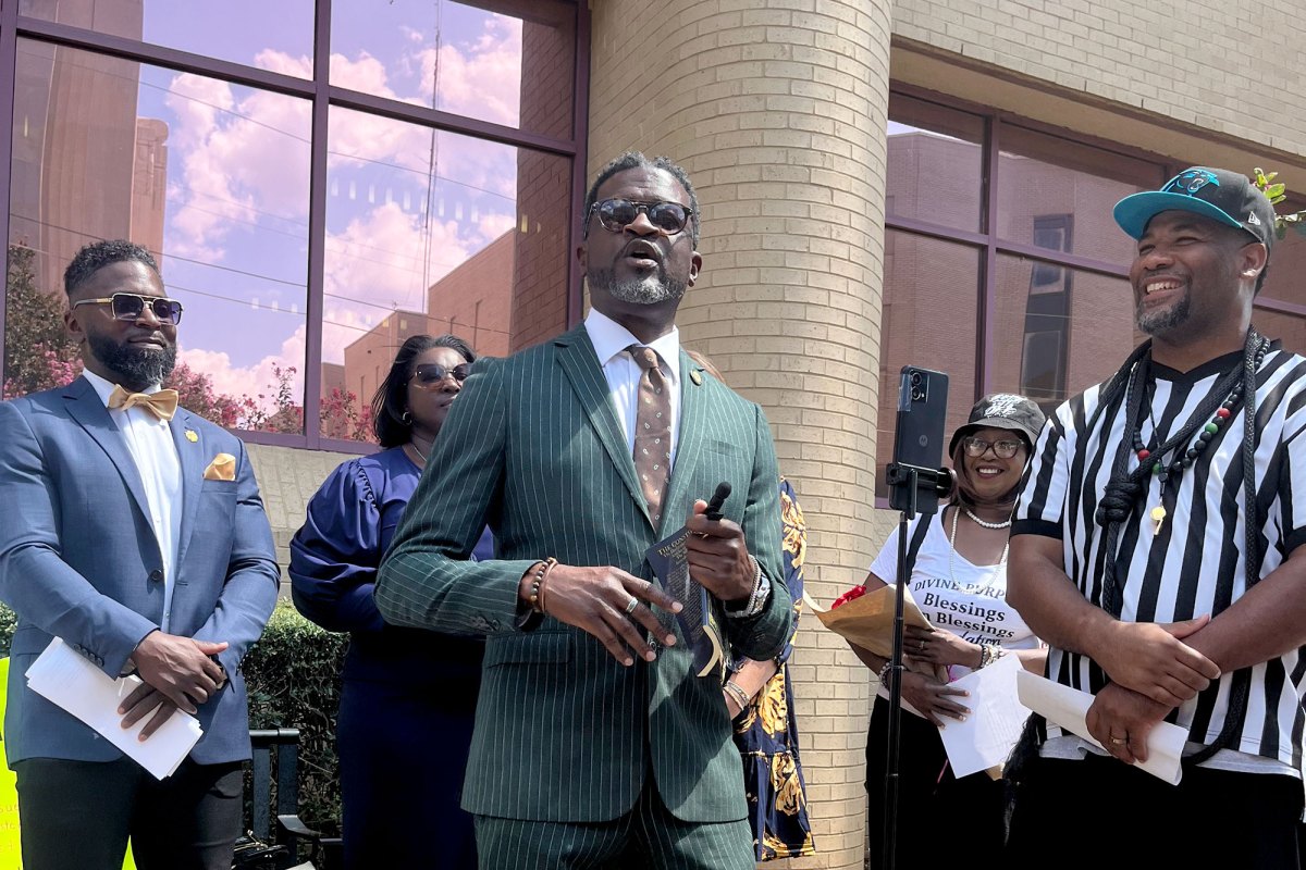 A man in a green pinstripped suit speaks outside of a brick building with pink reflective windows as others look on and smile