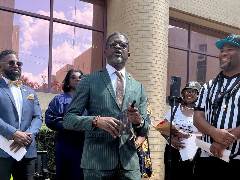 A man in a green pinstripped suit speaks outside of a brick building with pink reflective windows as others look on and smile