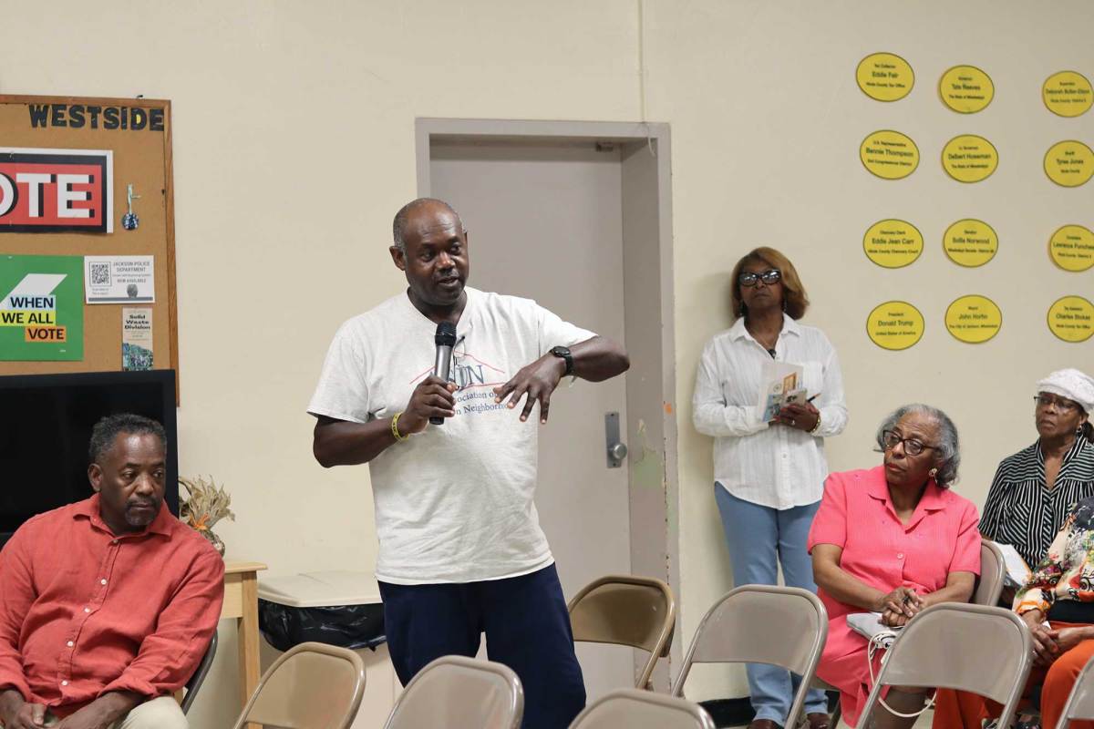 A man in a tshirt stands up in the seat row at a community meeting and talks, mic in hand, to the audience