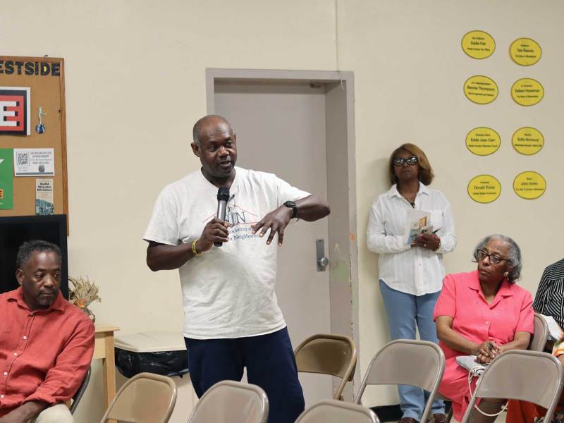 A man in a tshirt stands up in the seat row at a community meeting and talks, mic in hand, to the audience