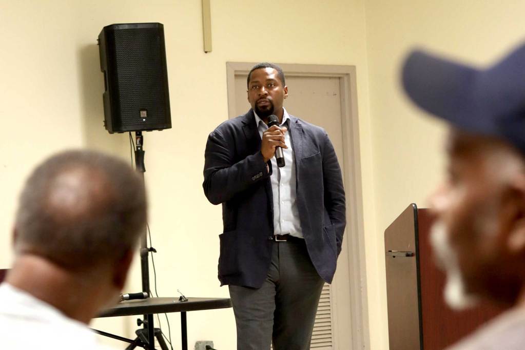 Ken Wilson stands in front of a community meeting and speaks, mic in hand