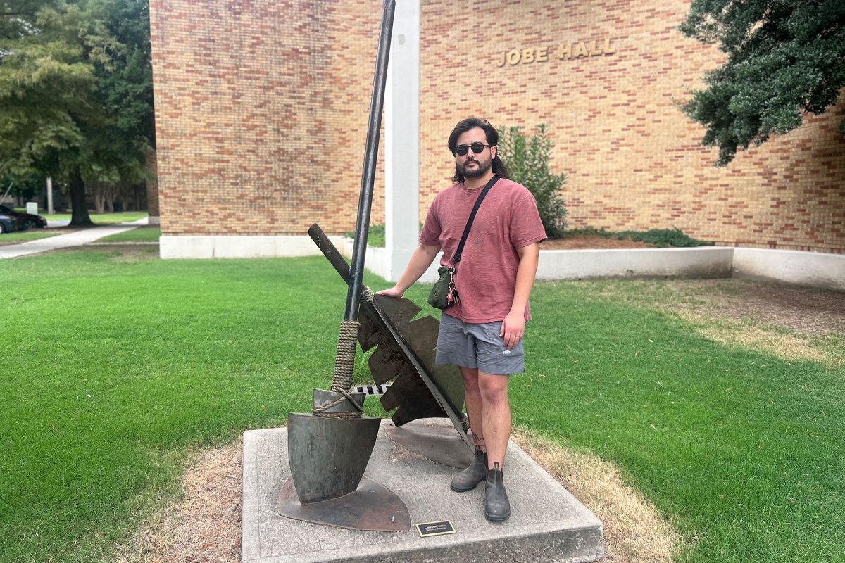 A man stands next to an art installation of a broken arrow outside of a brick building labeled 'Jobe Hall'