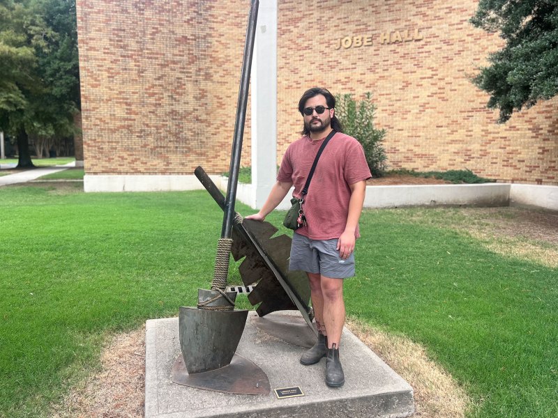 A man stands next to an art installation of a broken arrow outside of a brick building labeled 'Jobe Hall'
