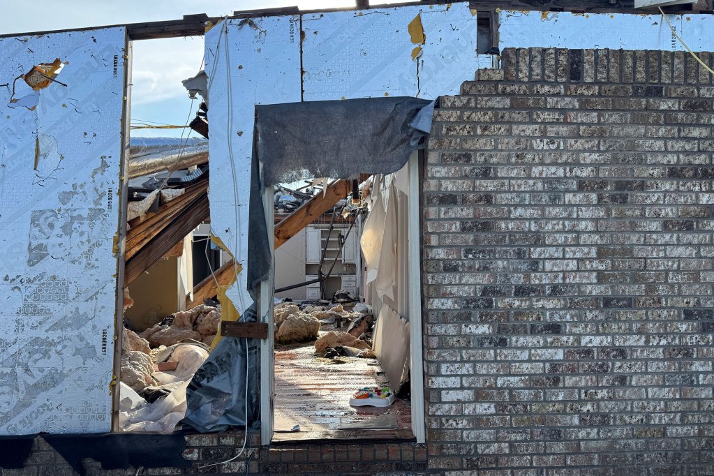An exterior view into a tornado damaged home, brick wall partially standing and door missing