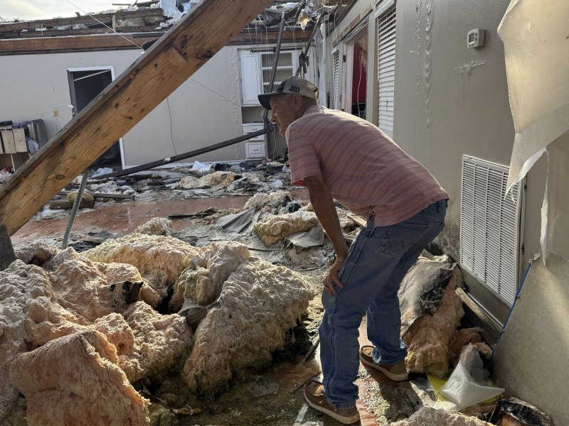 A man in a striped shirt and jeans surveys what's left of a tornado destroyed home