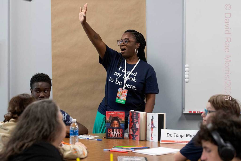 A woman stands in front of a panel, arm in air, with books standing before her