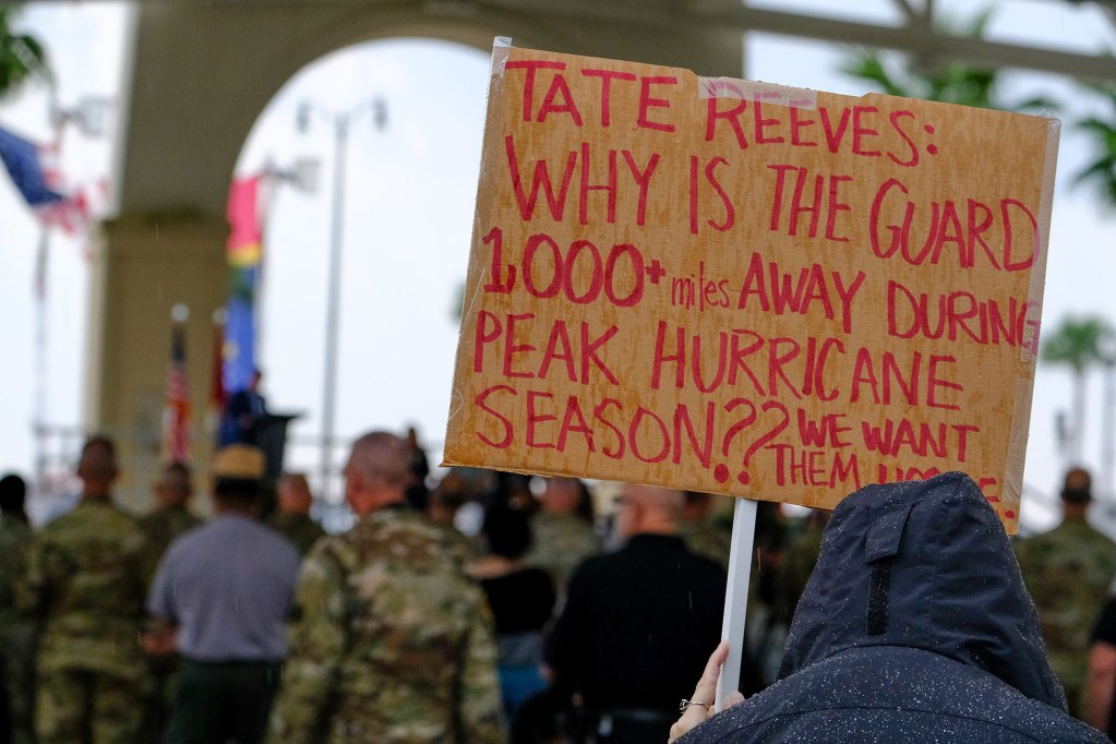 A person holds a sign in rain that reads 'Tate Reeves: Why is the Guard 1,000+ miles away during peak hurricane season?? We want them home.'