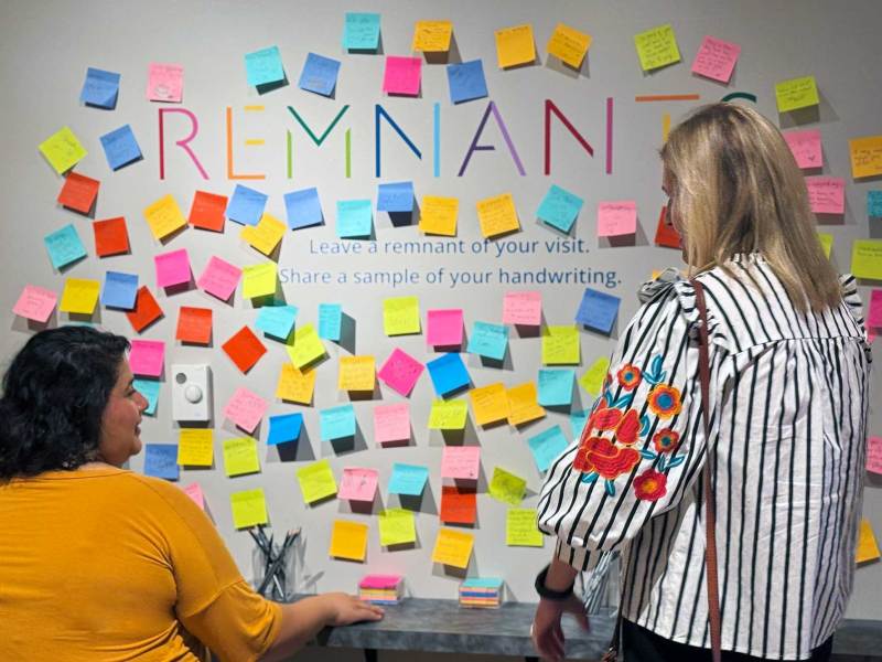 Two women add colorful sticky notes to a wall that reads REMNANTS in rainbow colors