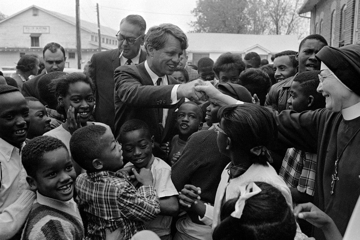 A black and white photo of Robert Kennedy shaking hands with a nun as many children surround them