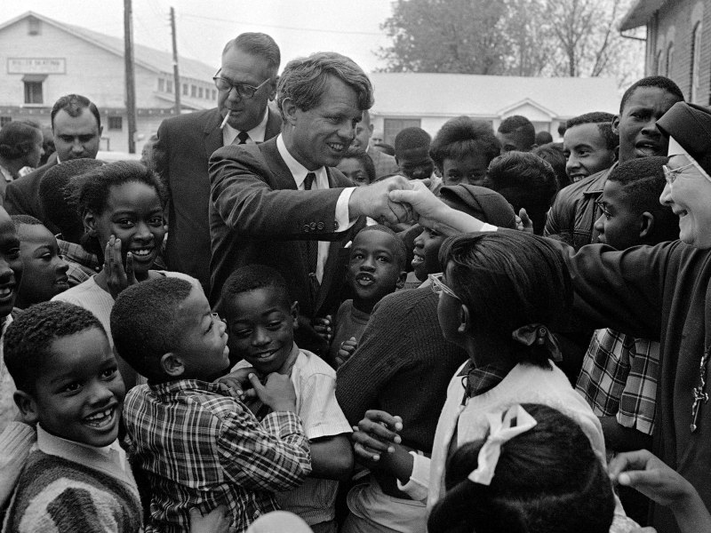 A black and white photo of Robert Kennedy shaking hands with a nun as many children surround them