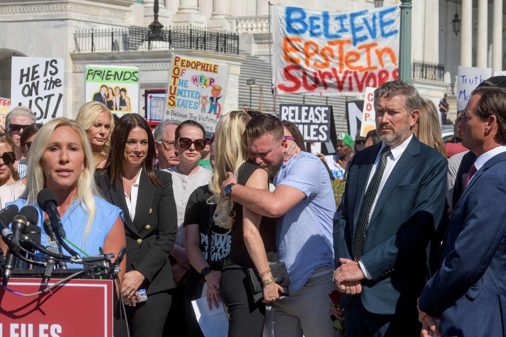 People embrace at a press conference outside, a sign behind them reads 'Believe Epstein Survivors'