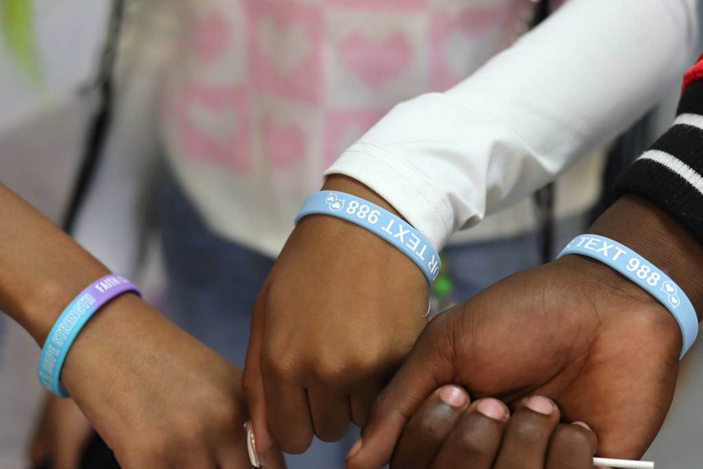 Three hands together all wearing blue plastic bracelets that read 'TEXT 988'
