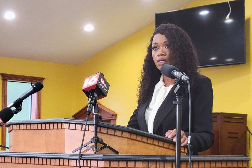 A woman speaking at a press conference held in a church with yellow walls