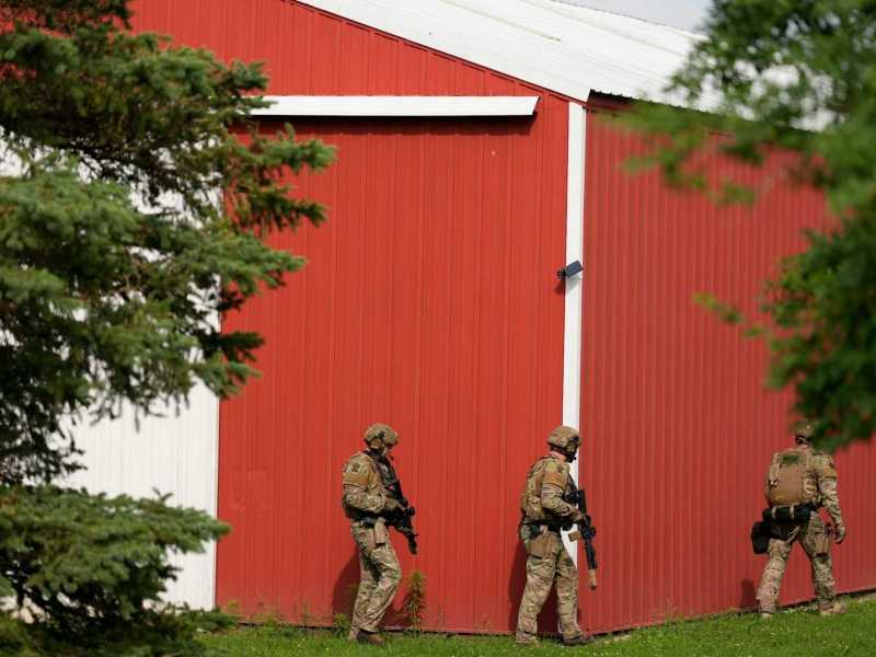 Armed military walk around the outside of a red barn with guns in ha