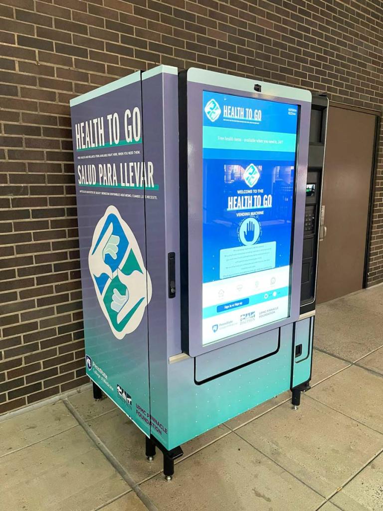 A teal and grey vending machine that says 'Health To Go' with large digital touchscreen on front, in front of a brick wall.