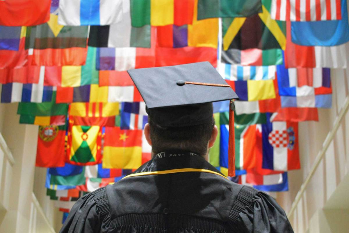 A view of a graduate in black robes and cap standing in a hallway, the ceiling covered in multiple countries flags