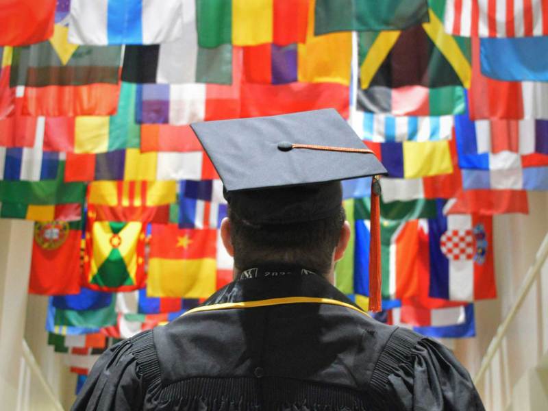 A view of a graduate in black robes and cap standing in a hallway, the ceiling covered in multiple countries flags