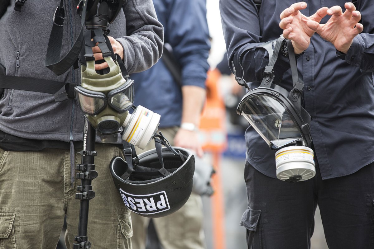A closeup of two people standing together in a group, holding gas masks. The person on the left is also holding camera equipment and a hardhat that reads PRESS in their hands