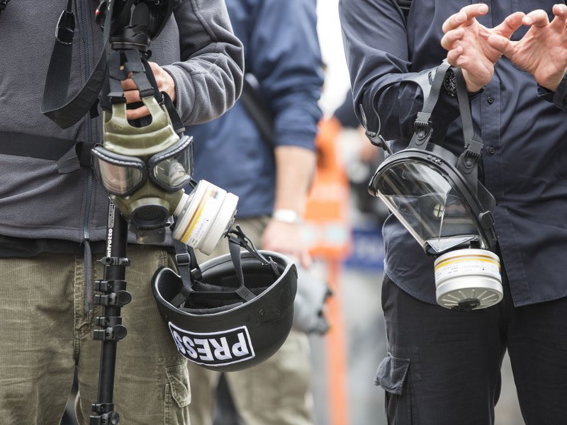 A closeup of two people standing together in a group, holding gas masks. The person on the left is also holding camera equipment and a hardhat that reads PRESS in their hands