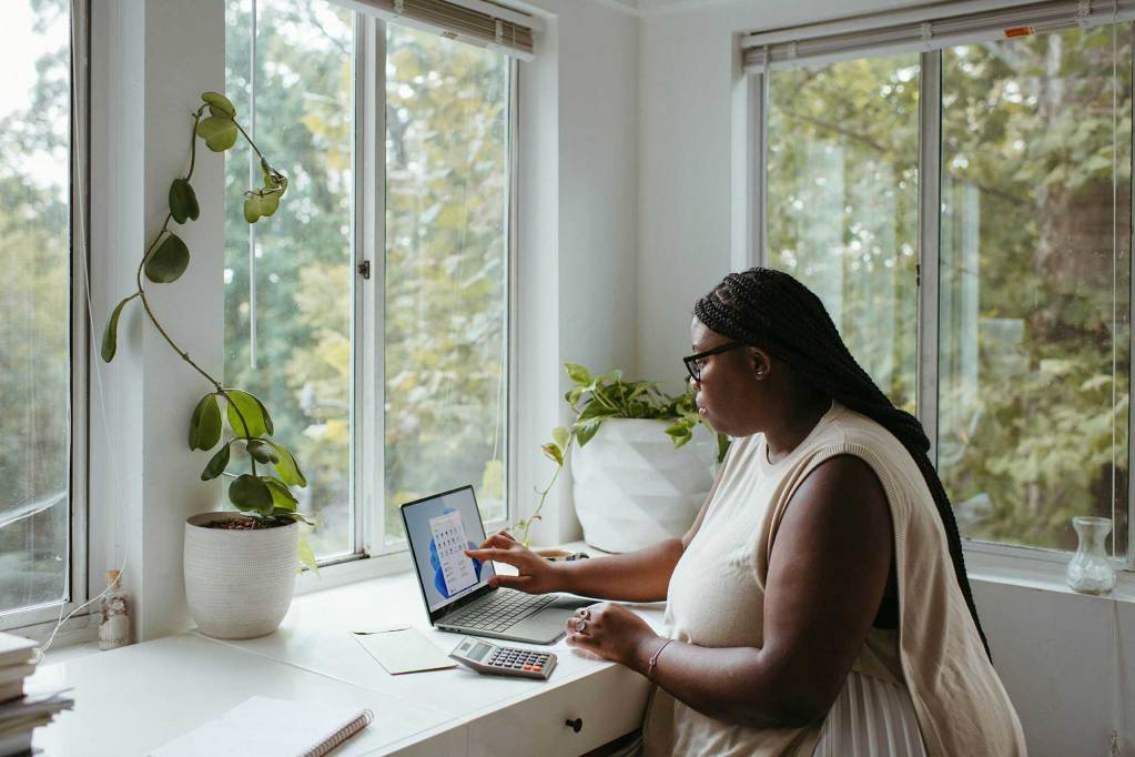 A woman sits at her laptop in a well lit white room with windows showing a green outside