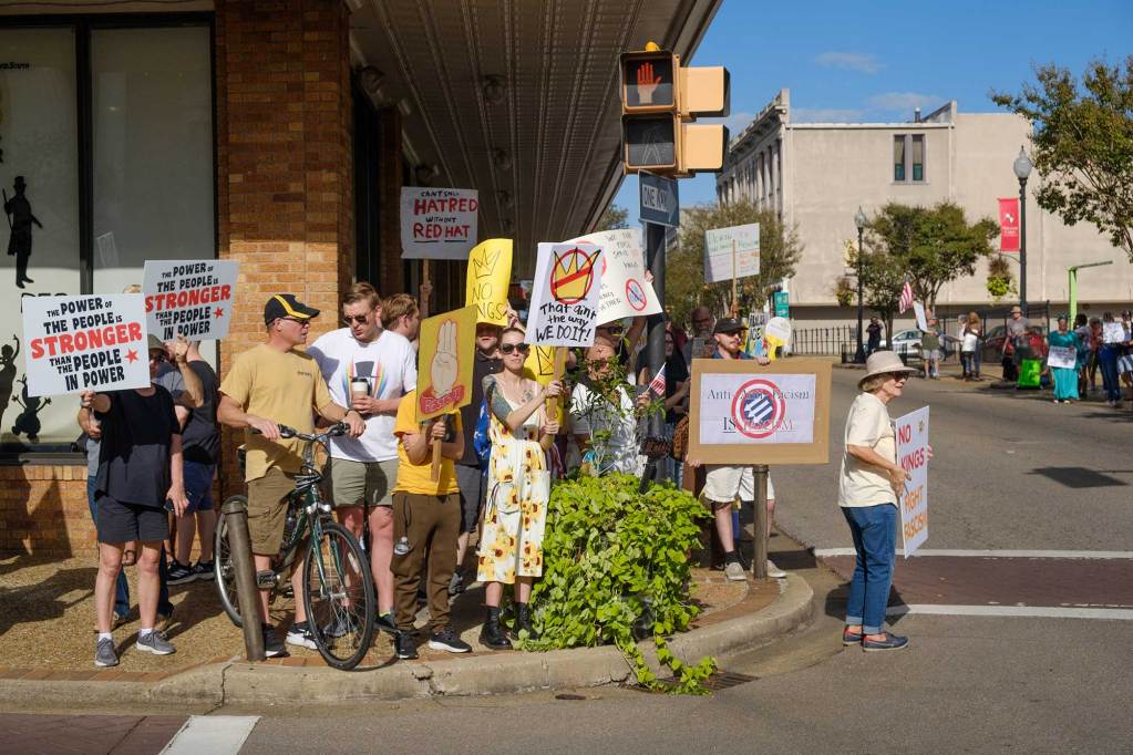 A group of protesters hold up signs on the sidewalk of a road. One reads 'The Power of the People is Stronger than the People in Power'