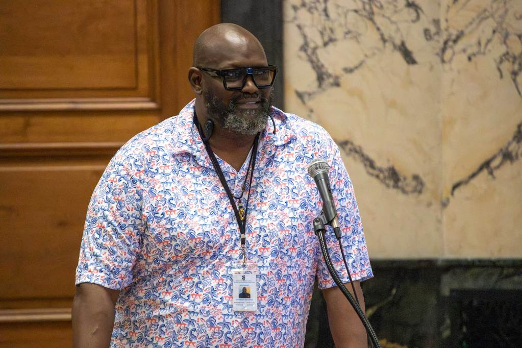 A bearded man in glasses and nautical print shirt speaks in a board meeting