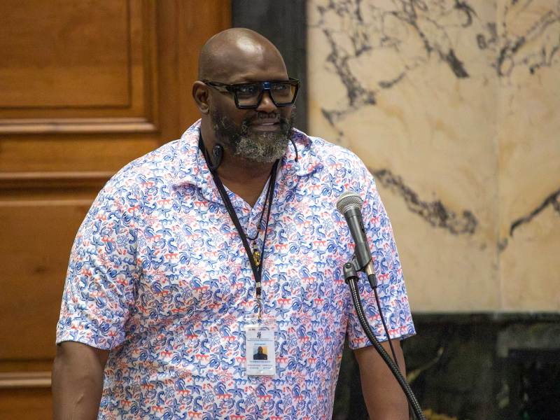 A bearded man in glasses and nautical print shirt speaks in a board meeting