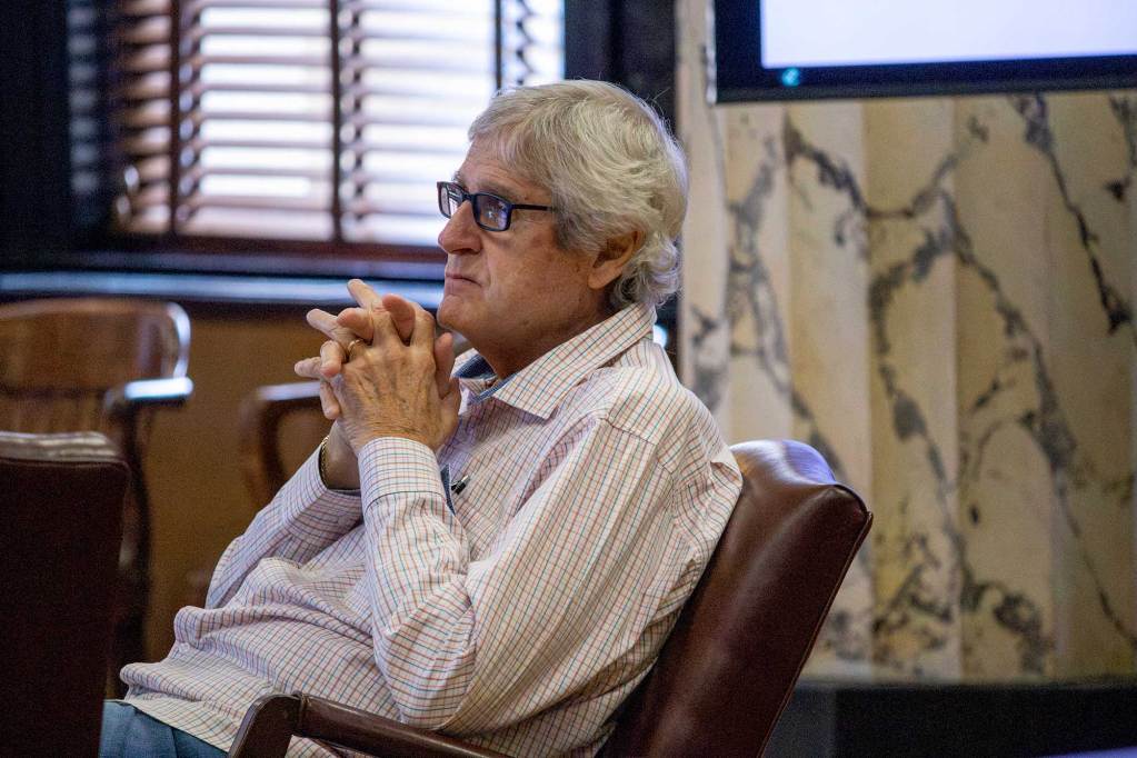 A man in glasses sits in a chair in a meeting room, hands steepled