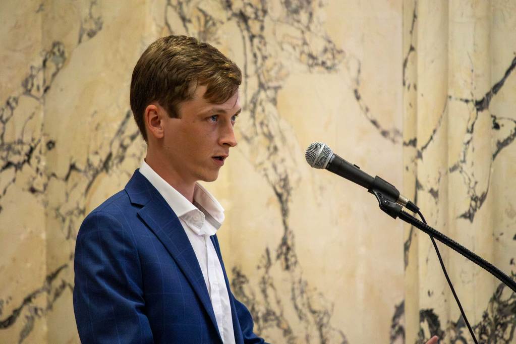 A man speaks at a microphone in front of a marble wall