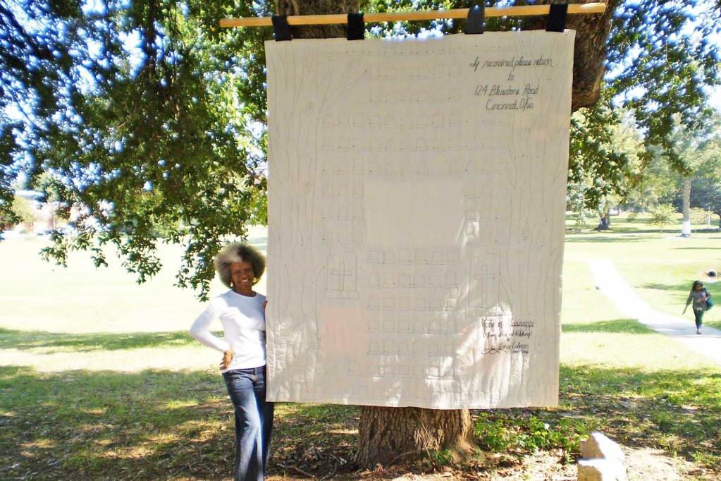 A woman stands outside next to a white quilt hanging from a tree beside her