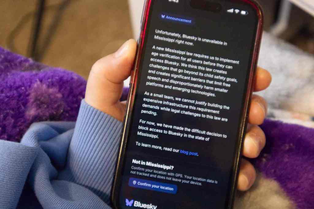 A hand holds a phone with a screen that shows the Bluesky blocked in Mississippi announcement.