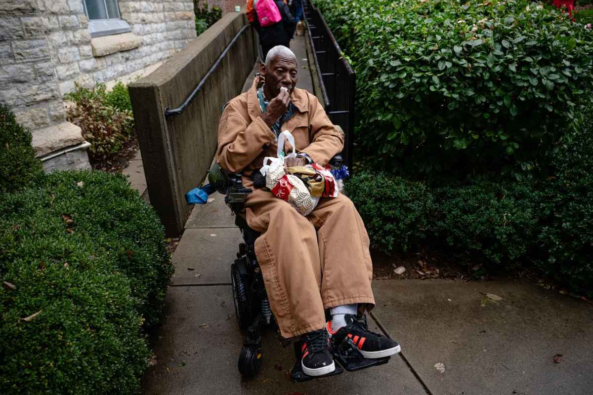 A Black man seated in a wheelchair. He is wearing a brown jacket and brown baggy pants and has a bag in his lap.