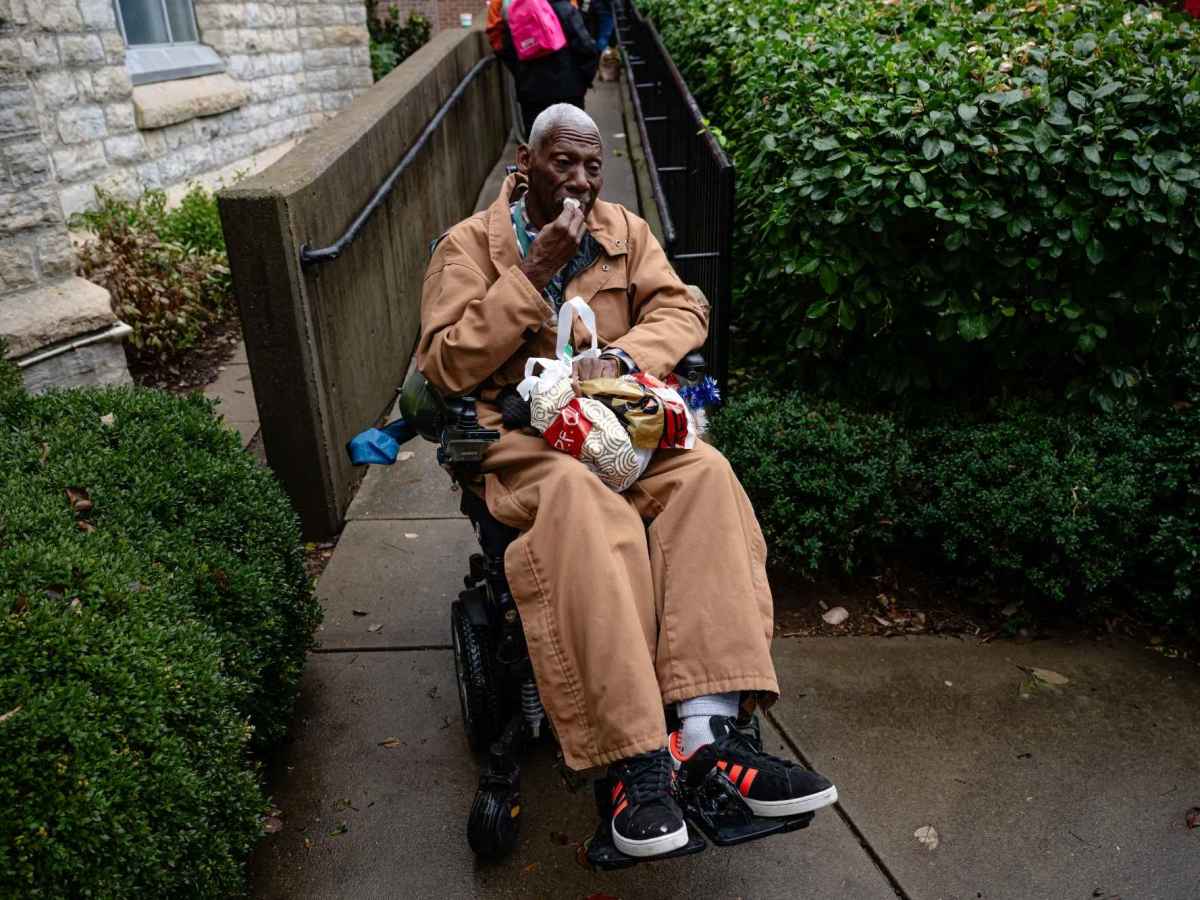 A Black man seated in a wheelchair. He is wearing a brown jacket and brown baggy pants and has a bag in his lap.