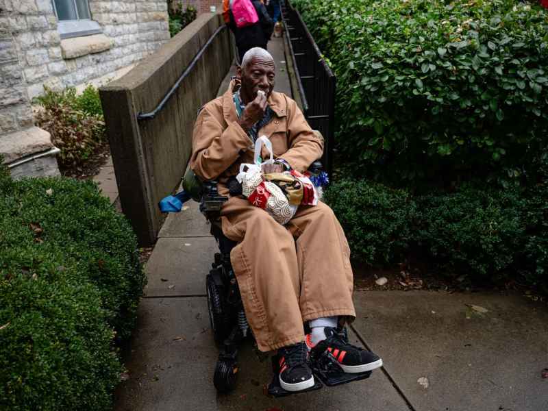 A Black man seated in a wheelchair. He is wearing a brown jacket and brown baggy pants and has a bag in his lap.