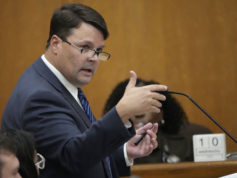 A man wearing a navy suit and black glasses gestures with his hands as he speaks into a microphone.