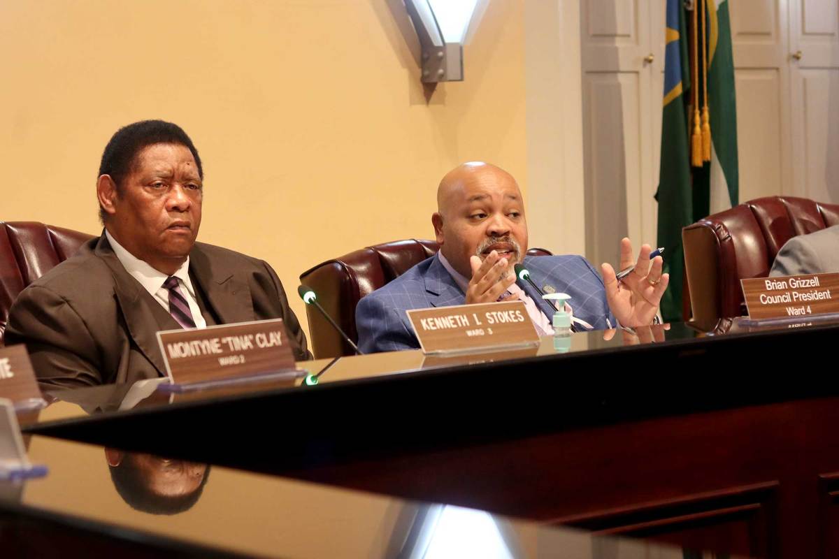Two councilmen, Kenneth Stokes and Brian Grizzell, sit in the council meeting room and discuss a topic with others