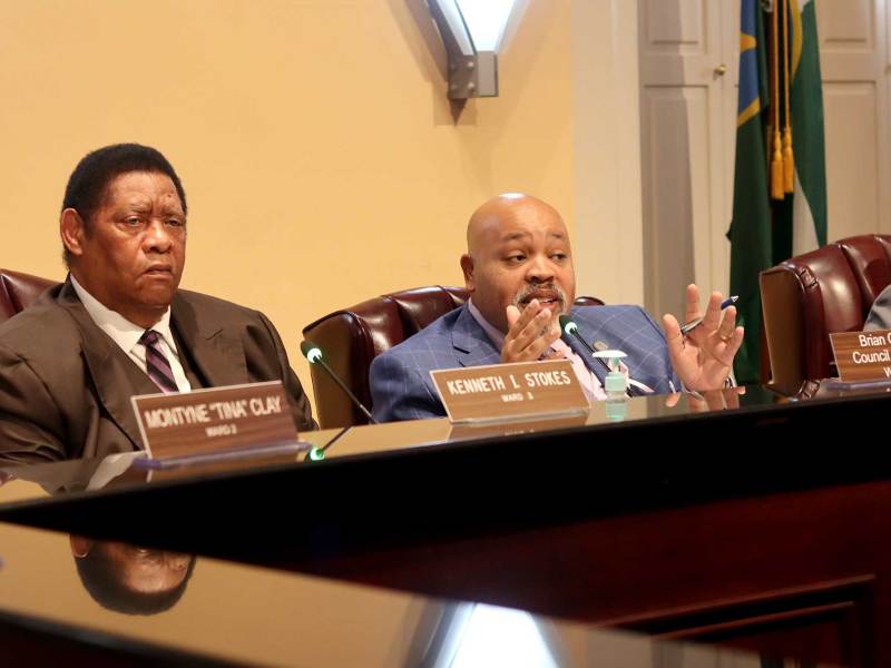 Two councilmen, Kenneth Stokes and Brian Grizzell, sit in the council meeting room and discuss a topic with others