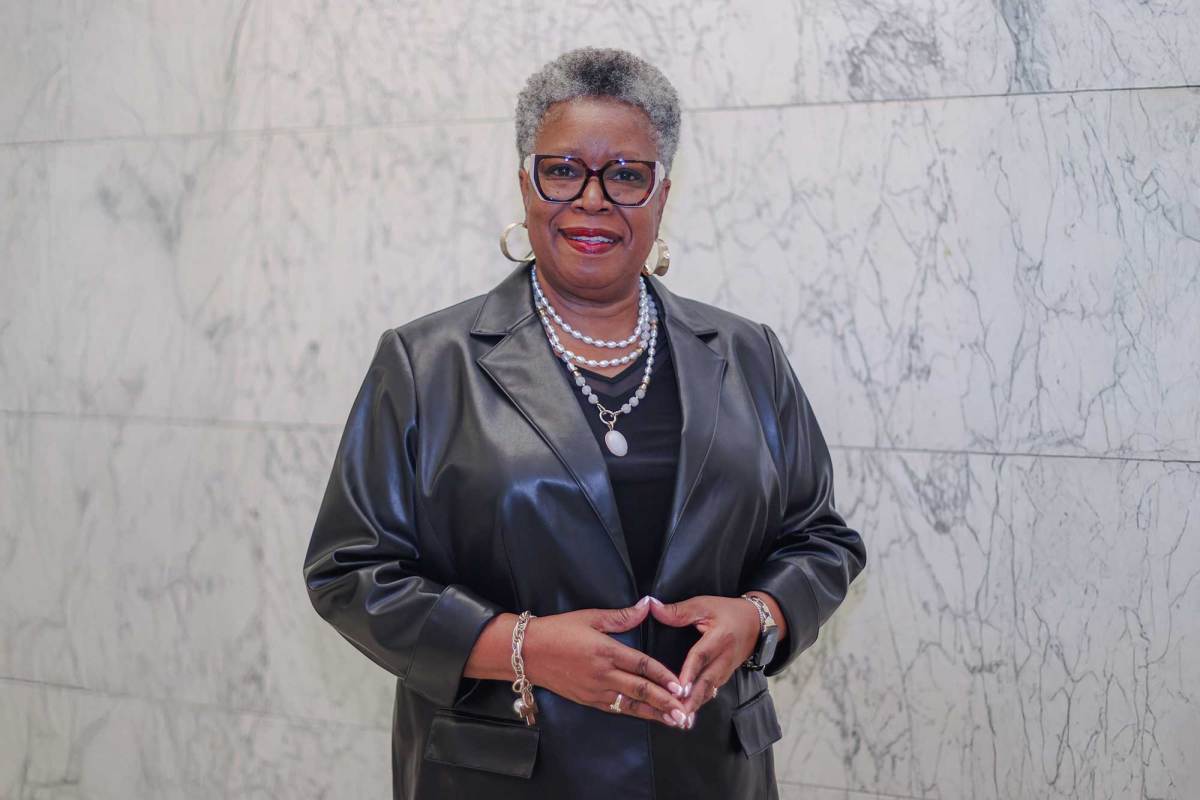 A woman wearing black framed glasses and a black blazer stands in a marble hallway with hands pressed together