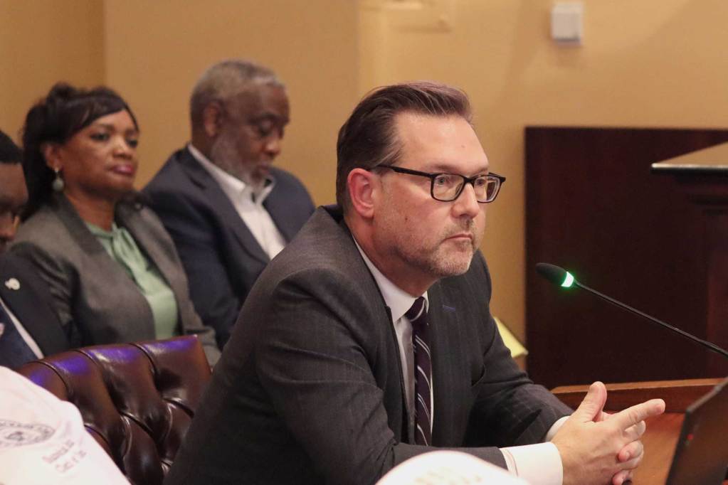 Drew Martin wearing a grey suit, seated and speaking at City Hall