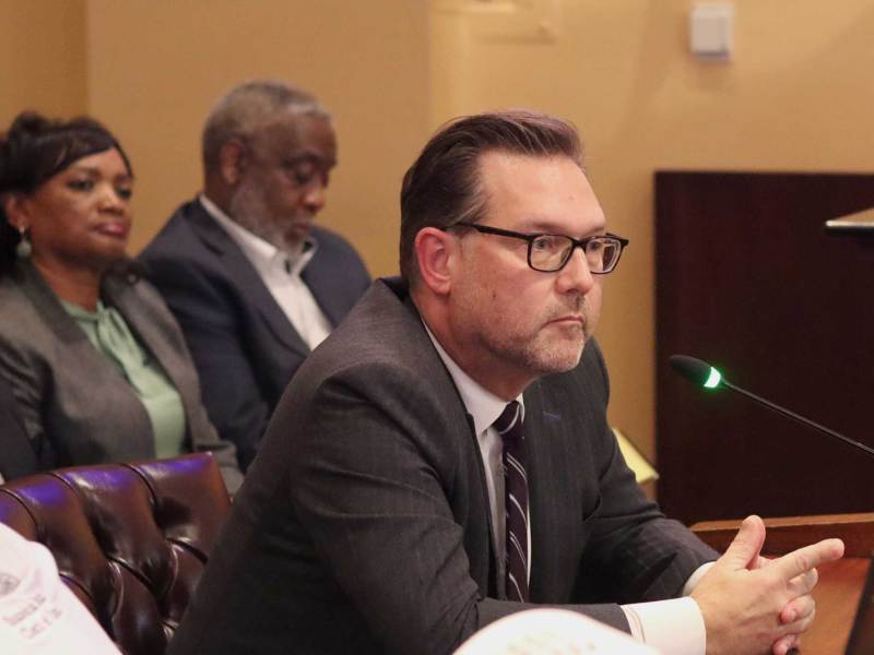 Drew Martin wearing a grey suit, seated and speaking at City Hall