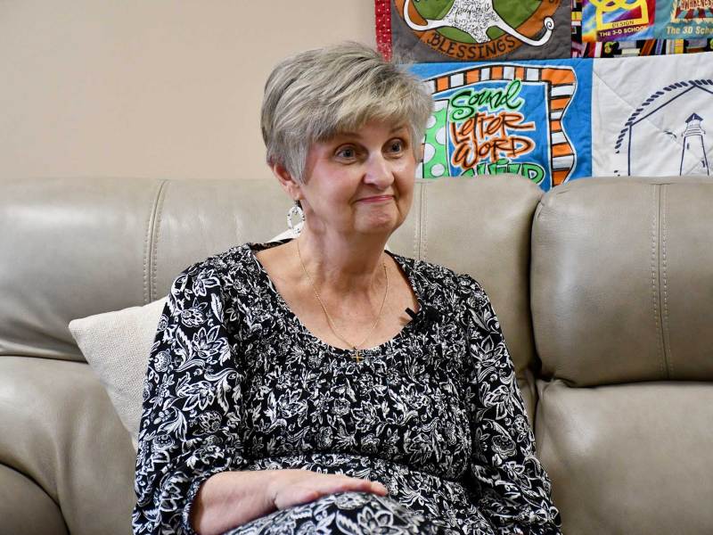 A woman with short hair wearing a black-and-white dress sits on a gray leather couch.