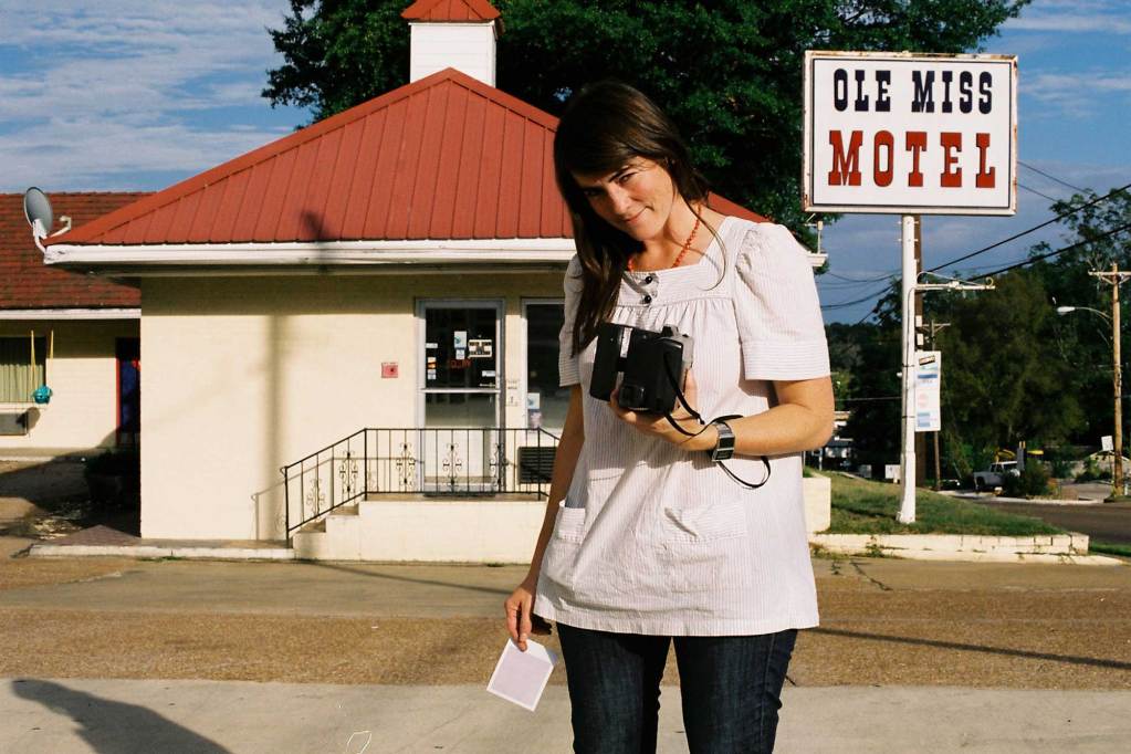 A woman takes self portrait with a camera held before her as she stands in front of the Ole Miss Motel