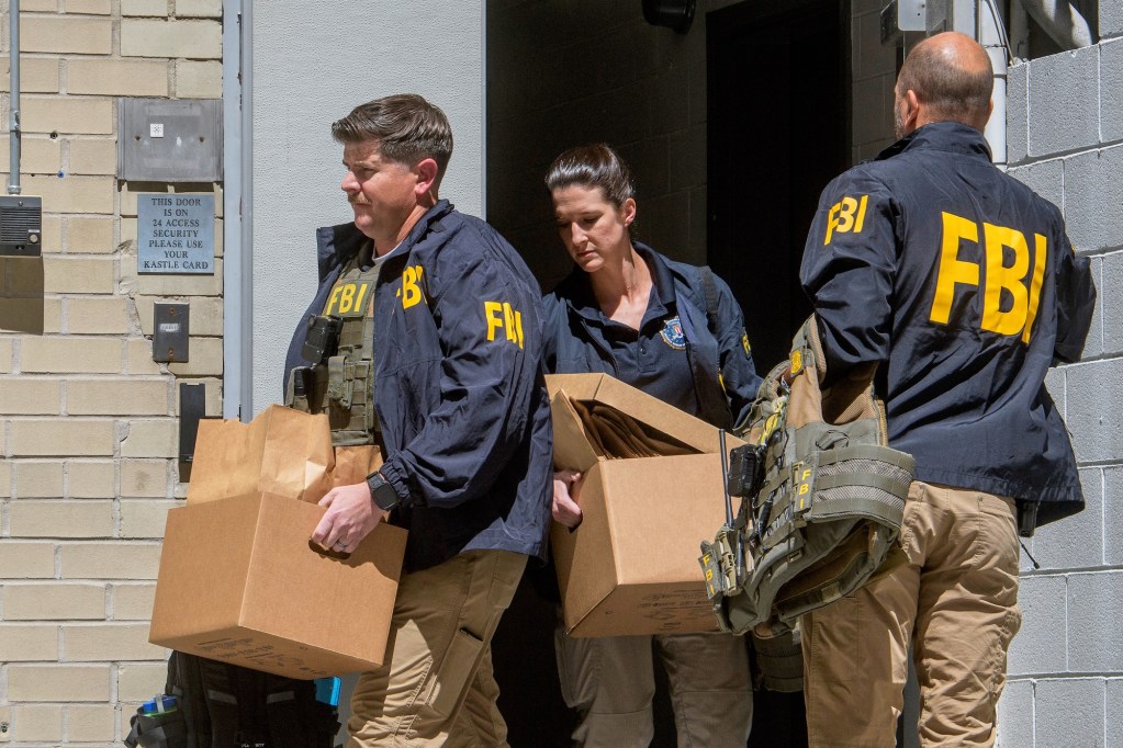 2 FBI agents carry boxes out of a building while another stands by